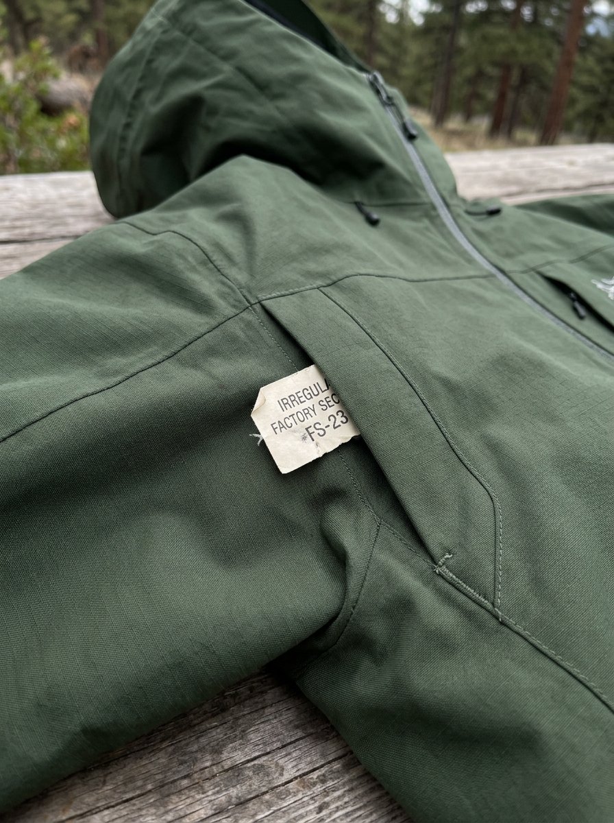 Close up of an olive green outdoor jacket featuring a paper tag labeled irregular factory second in a chest pocket.